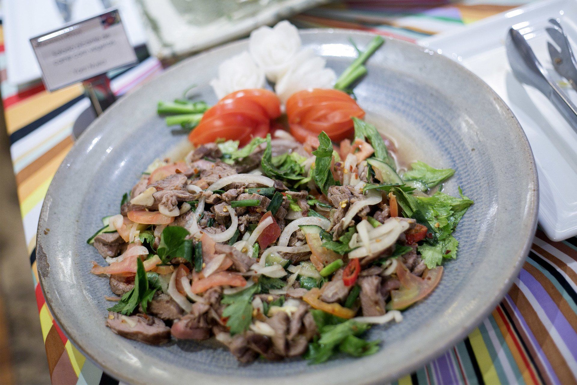 Salada com legumes e carne cozida em um prato cinza, com tomates e flores brancas ao lado.