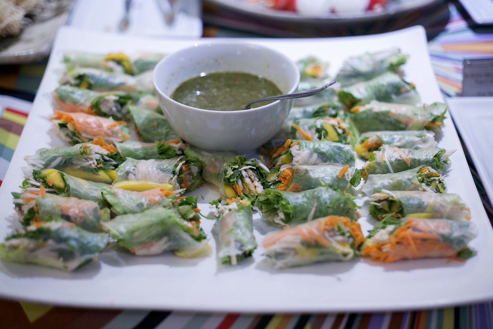 Prato de rolinhos primavera coloridos com uma tigela central de molho verde.