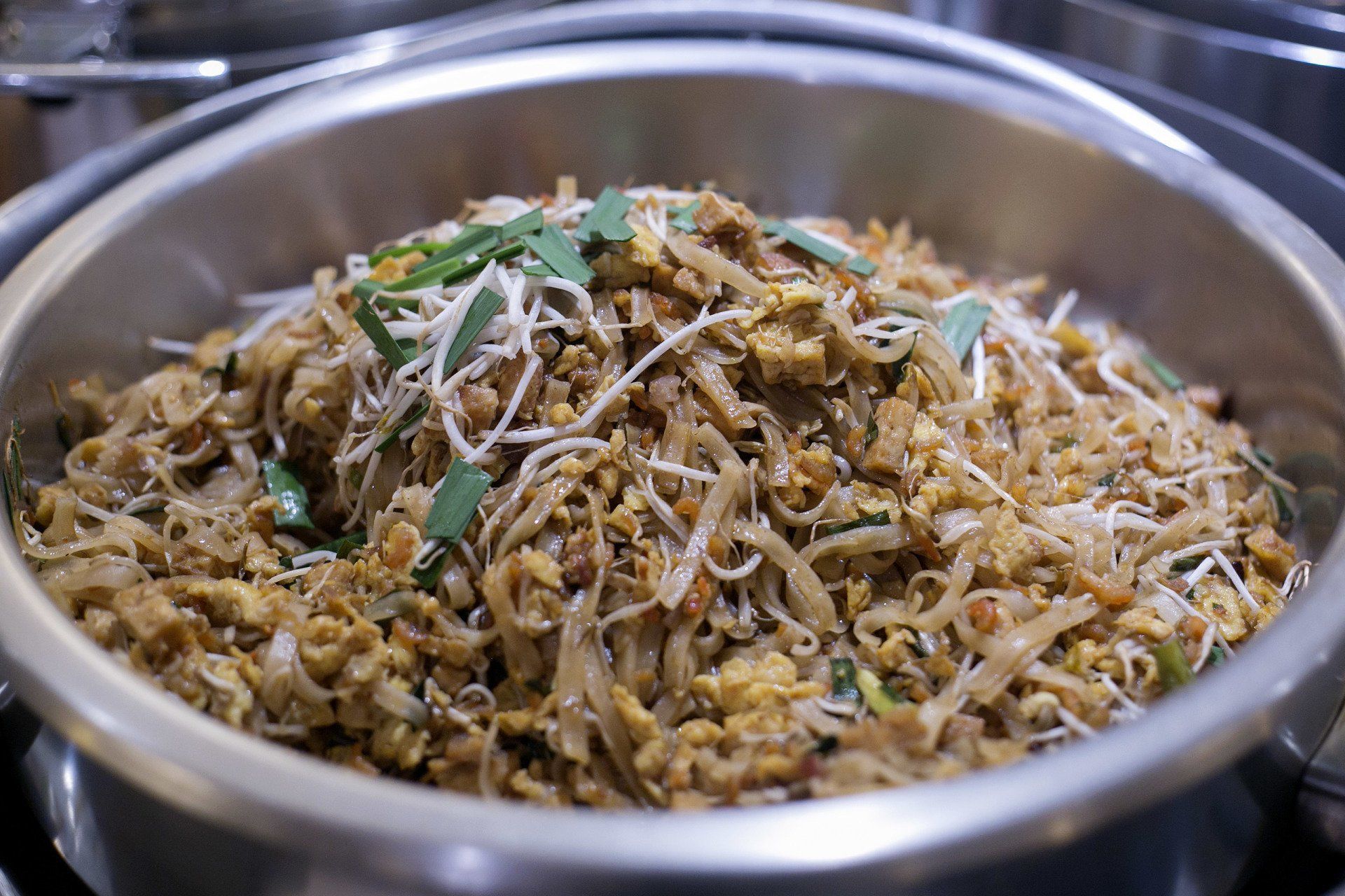 Macarrão Pad Thai em uma travessa de prata, decorado com brotos de feijão e ervas.