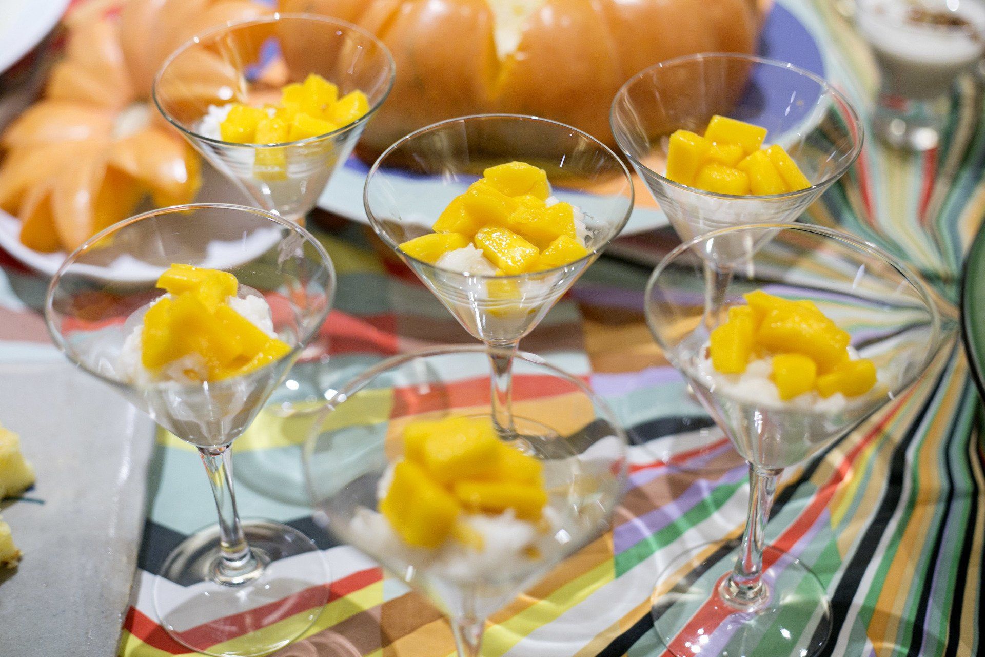 Arroz glutinoso com manga em taças de martini, toalha de mesa colorida e suco de laranja ao fundo.