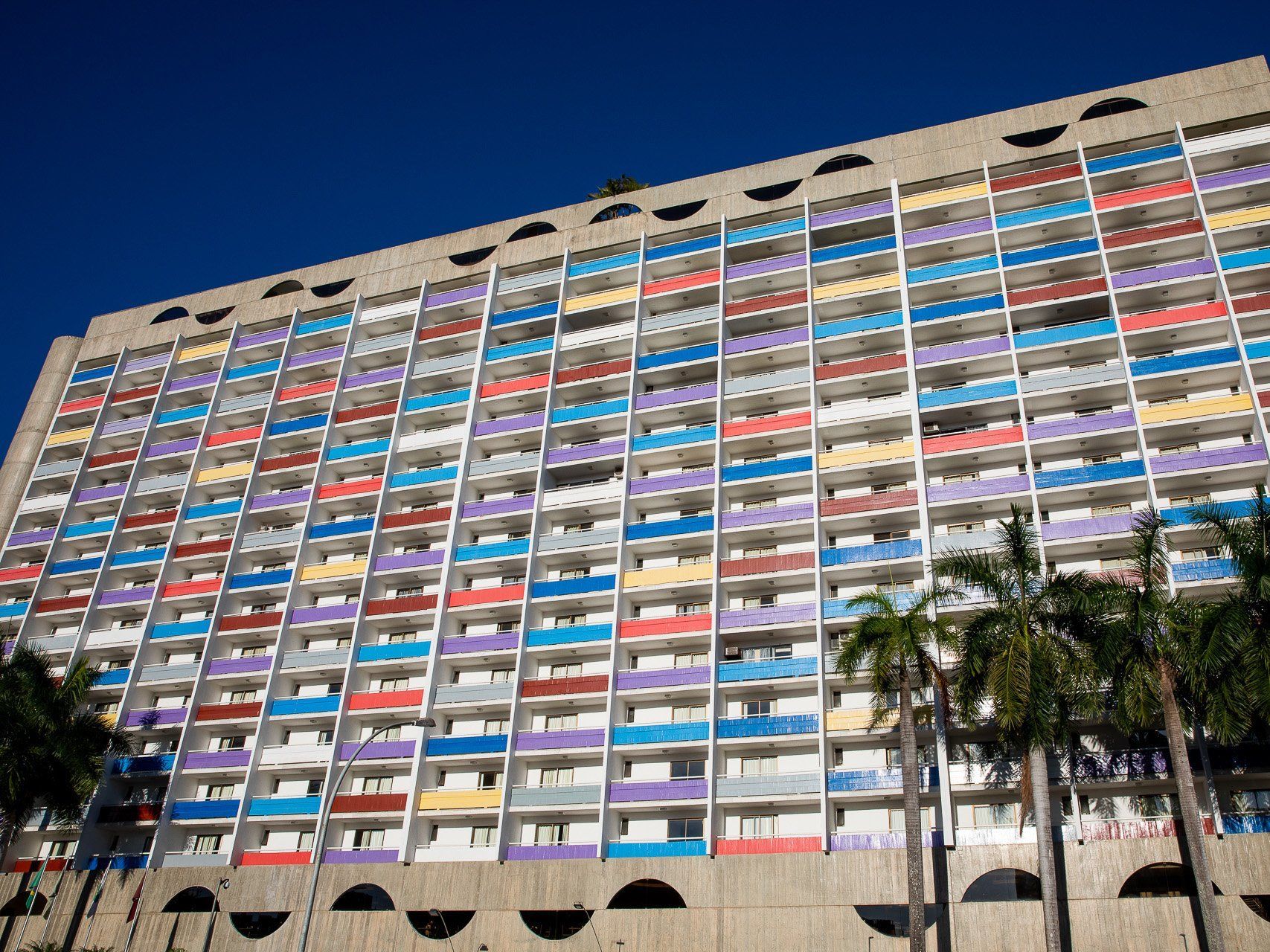 Um grande edifício com muitas varandas coloridas sob um céu azul claro, emoldurado por palmeiras.