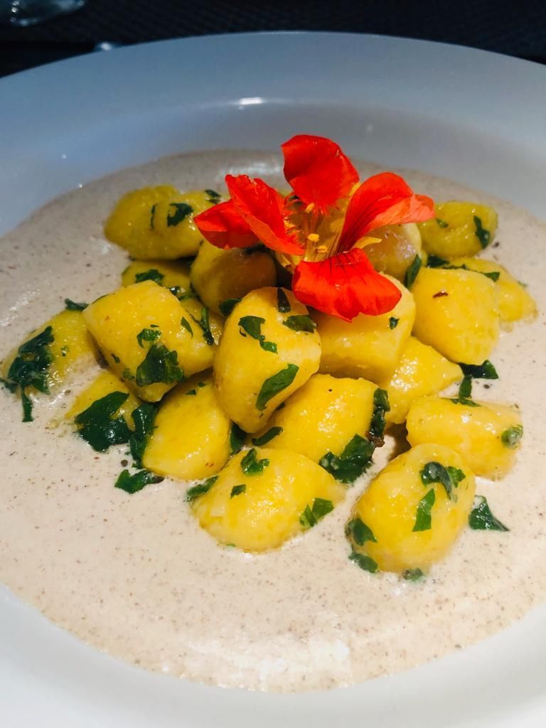 Nhoque amarelo com ervas verdes, guarnição de flores vermelhas e molho cremoso em uma tigela branca.