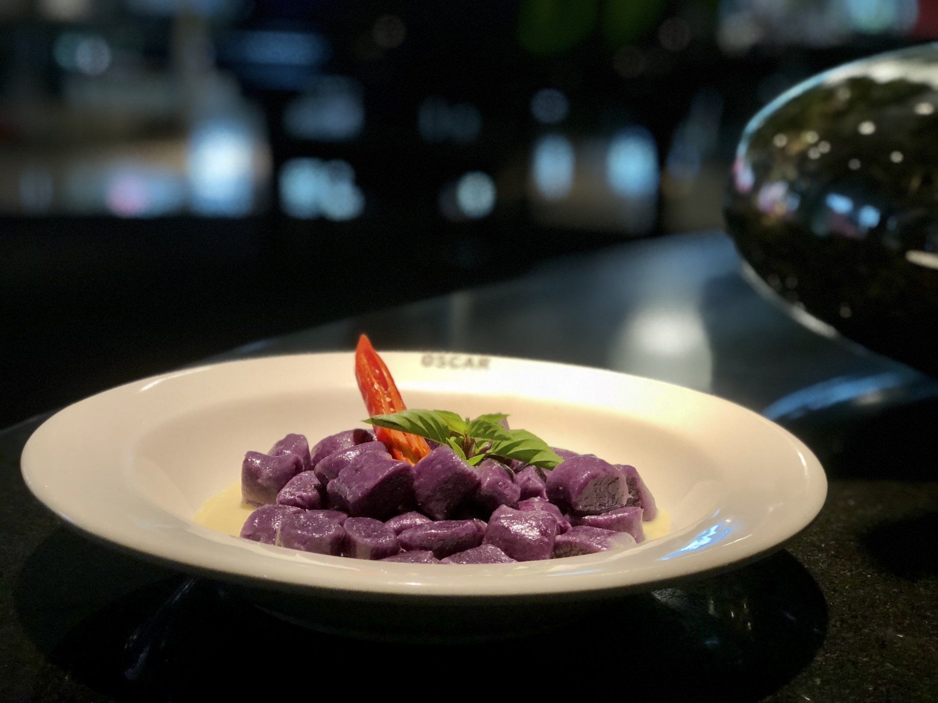 Nhoque roxo em um prato branco, decorado com manjericão e pimentão vermelho, em um balcão escuro.