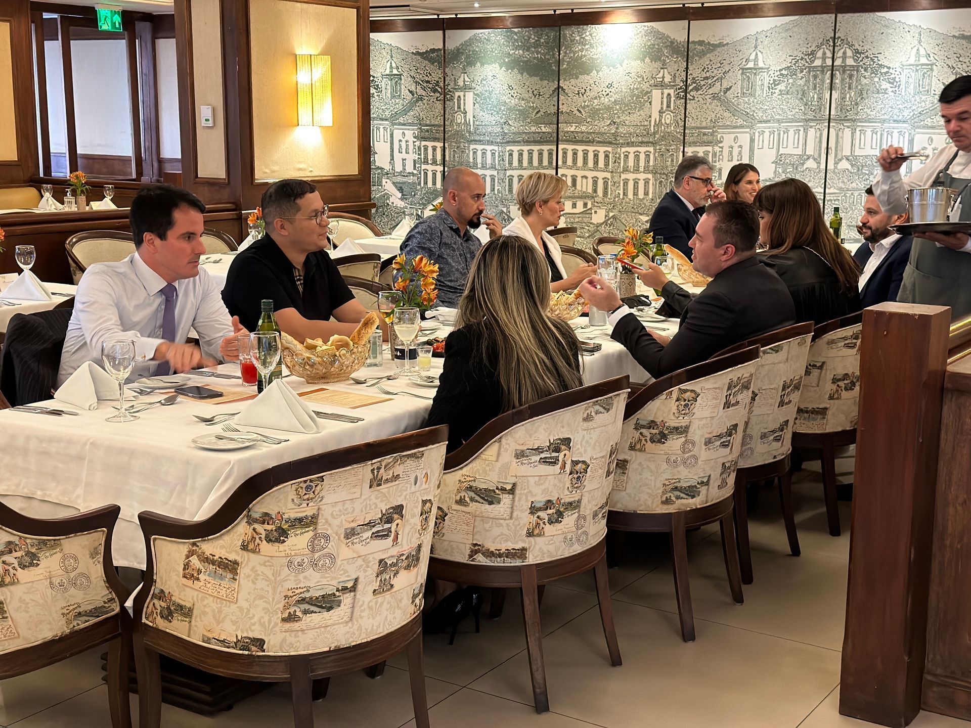 Grupo de pessoas sentadas em uma longa mesa em um restaurante, interagindo entre si e sendo servidas por um garçom.