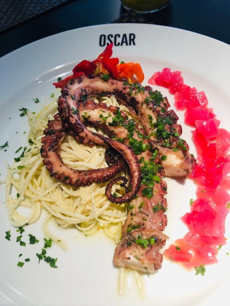 Polvo grelhado sobre espaguete, guarnecido com ervas picadas e cebolas vermelhas em conserva em um prato branco.