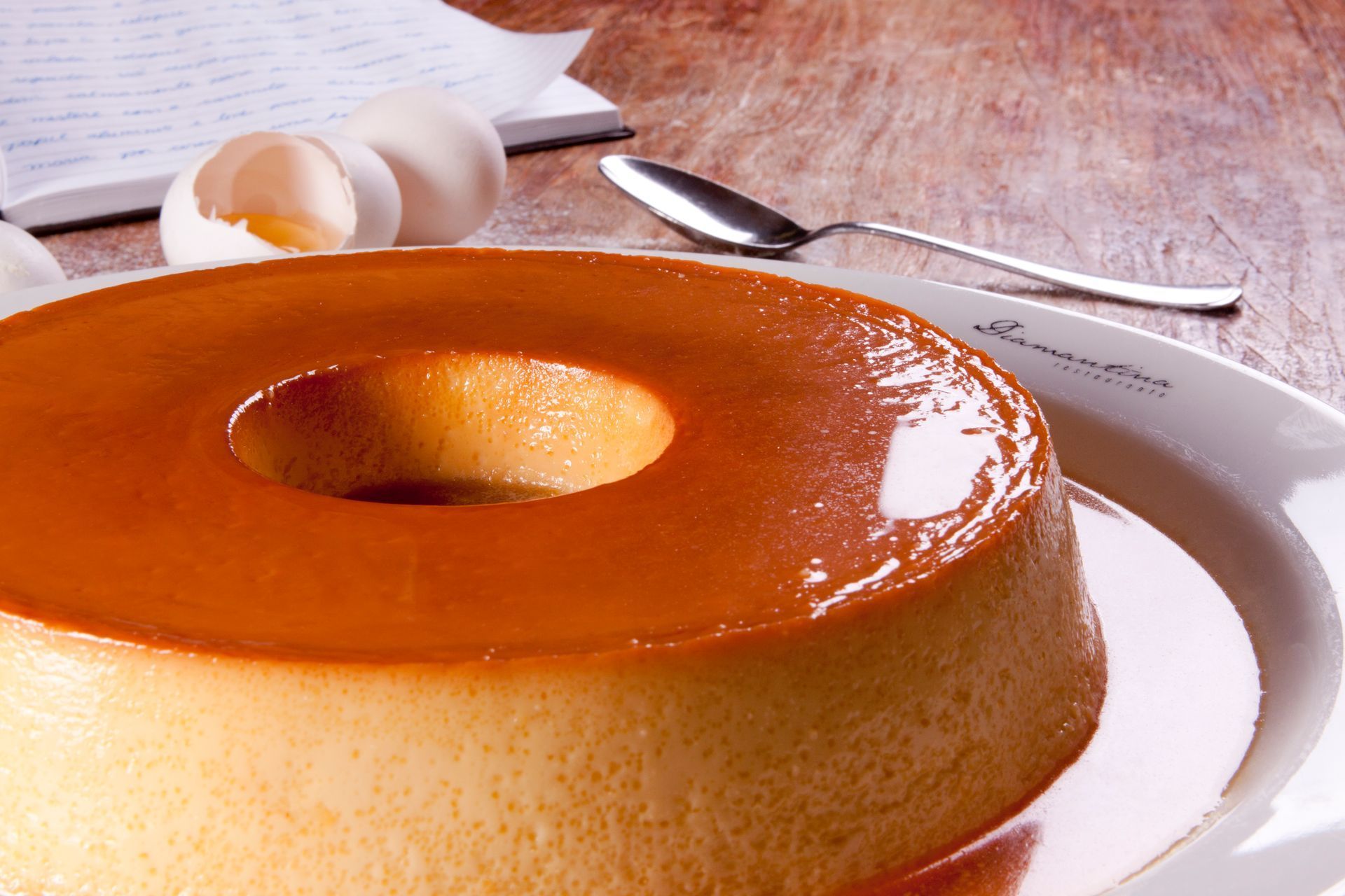 Uma sobremesa de flan dourada em um prato branco, com uma colher e cascas de ovos ao fundo.