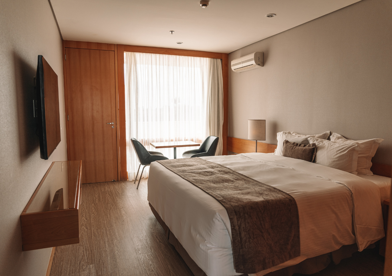 Quarto de hotel com uma cama, janela com cortinas, duas cadeiras e uma TV.