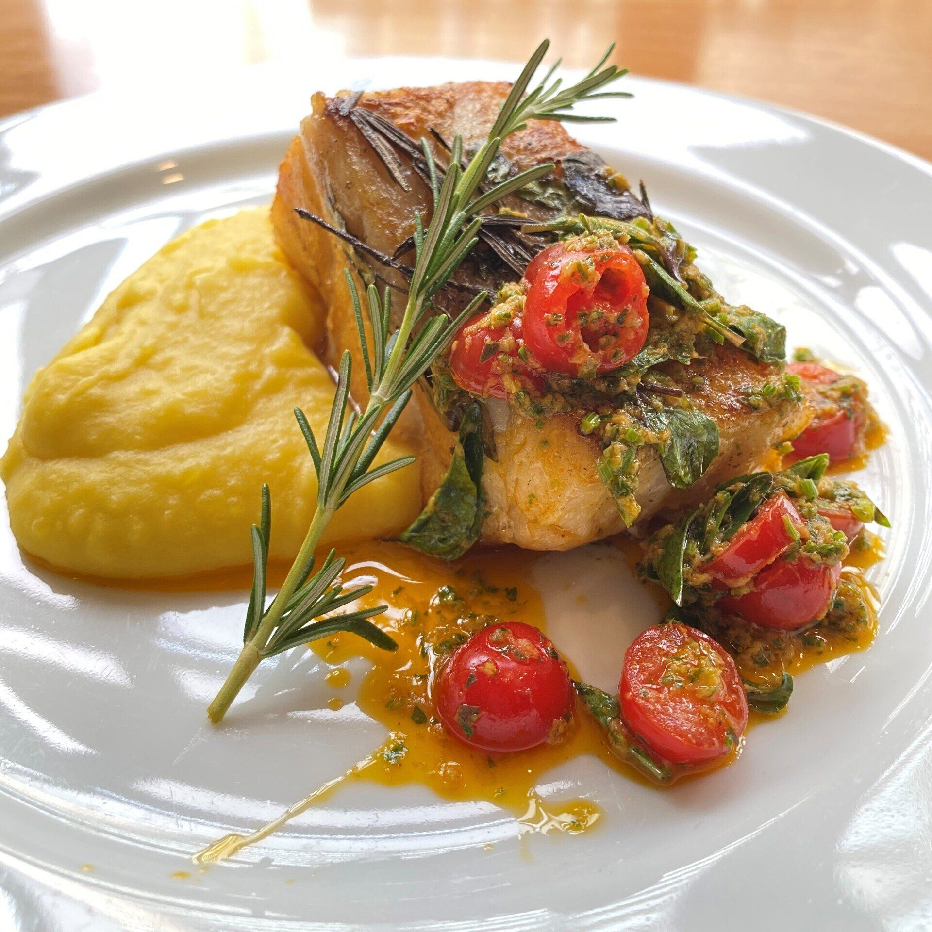 Peixe com molho de tomate e manjericão, purê de batatas amarelo e um raminho de alecrim em um prato branco.