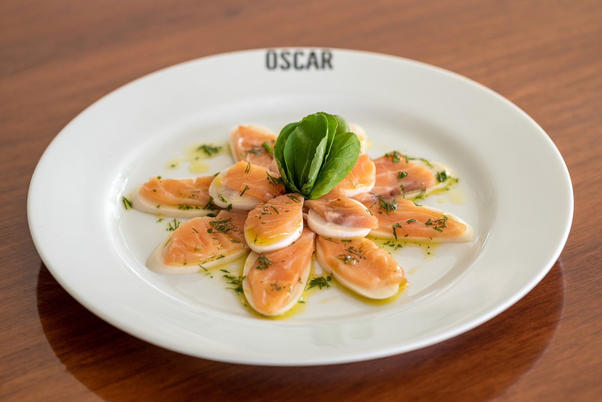 Carpaccio de salmão disposto em padrão floral, decorado com ervas, em um prato branco.