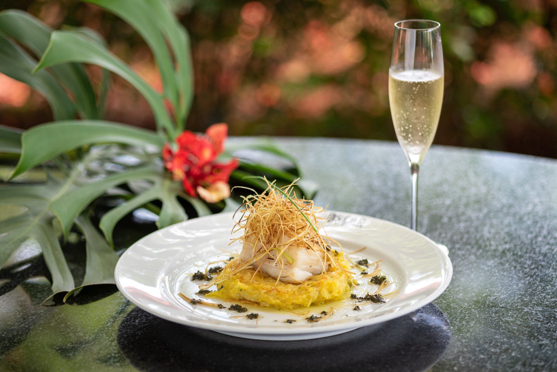 Prato de peixe com base amarela, coberto com guarnição crocante, ao lado de champanhe.