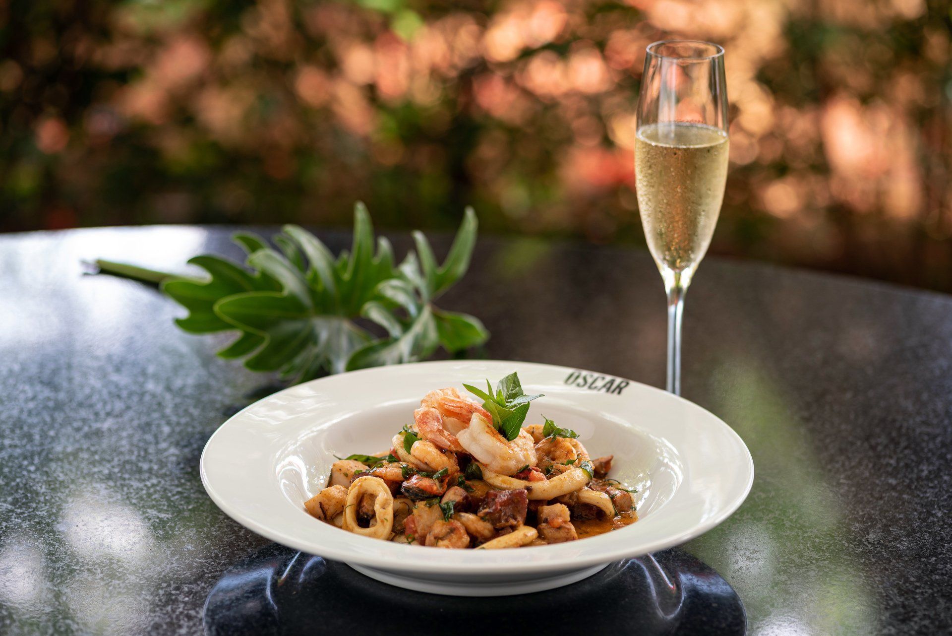 Prato de frutos do mar e champanhe na mesa, com um raminho de folhas verdes, ao ar livre.
