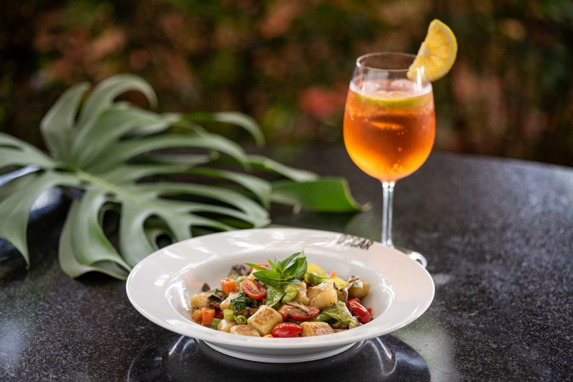 Prato de nhoque com tomate e manjericão, servido com um coquetel decorado com uma rodela de limão, ao ar livre.