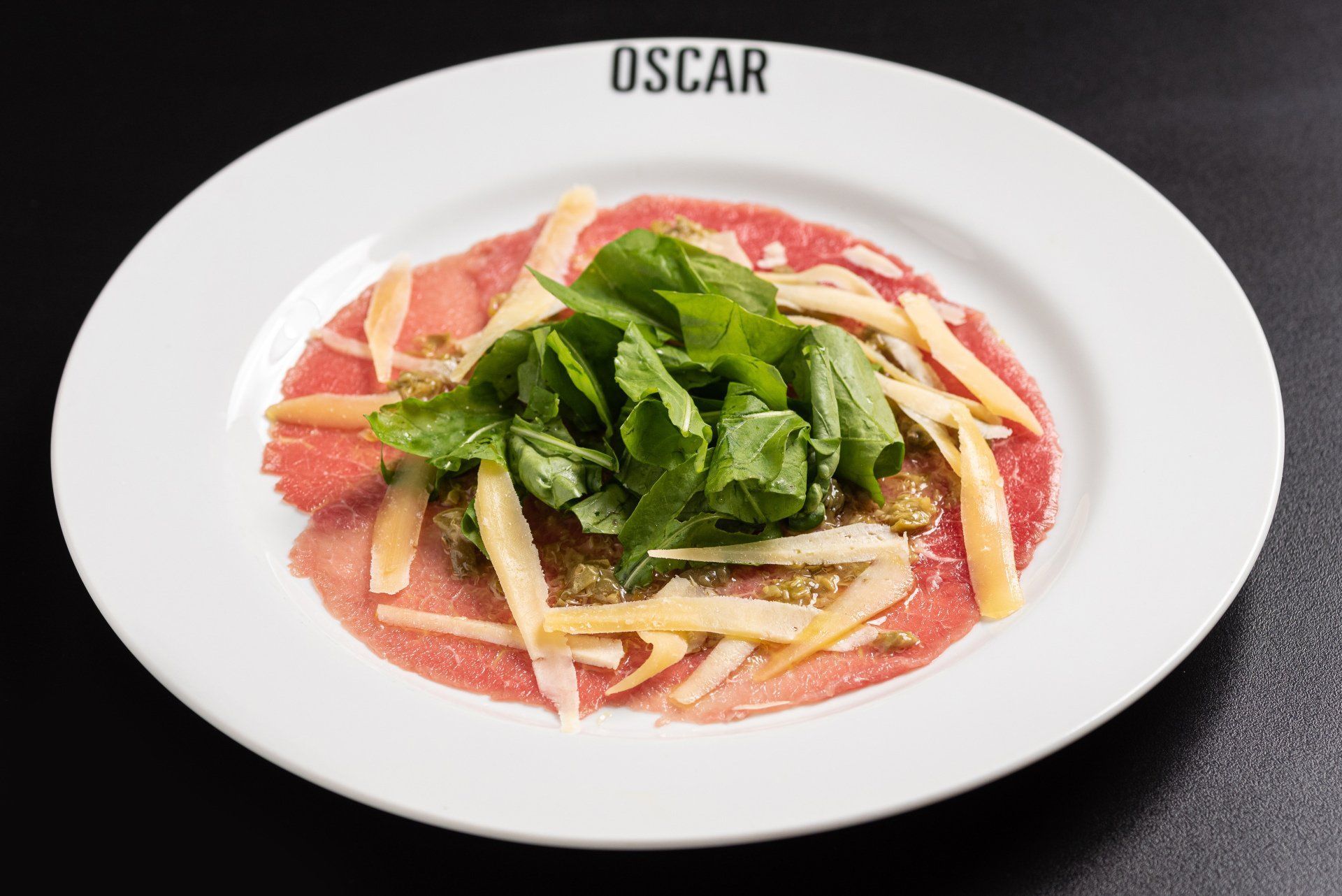 Carpaccio em um prato branco, decorado com rúcula e queijo. Logotipo do restaurante Oscar ao fundo.