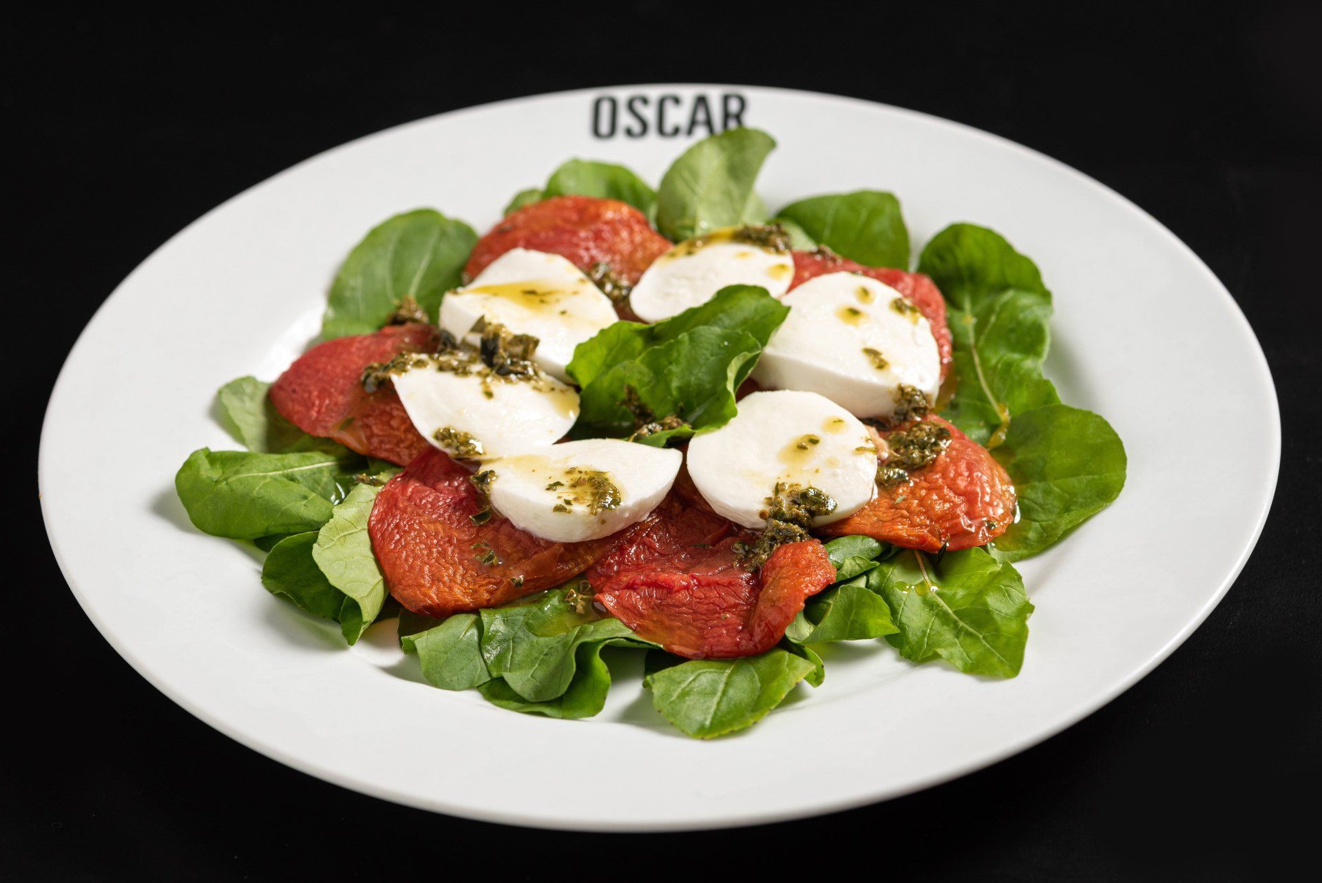 Salada com mussarela, tomates vermelhos, pesto e verduras em um prato branco, com o logotipo do 