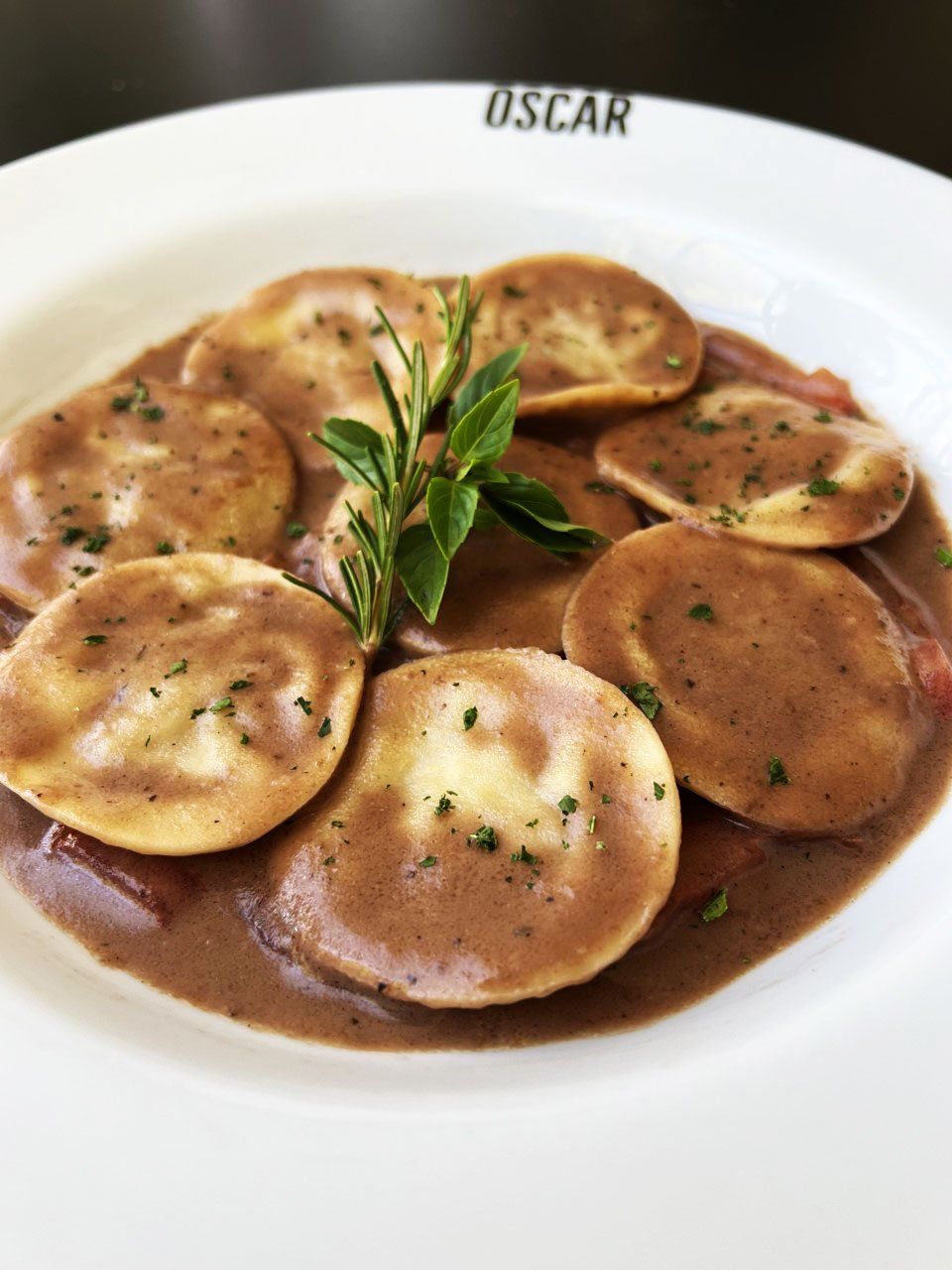 Ravioli ao molho marrom, decorado com ervas, em um prato branco. O prato tem 