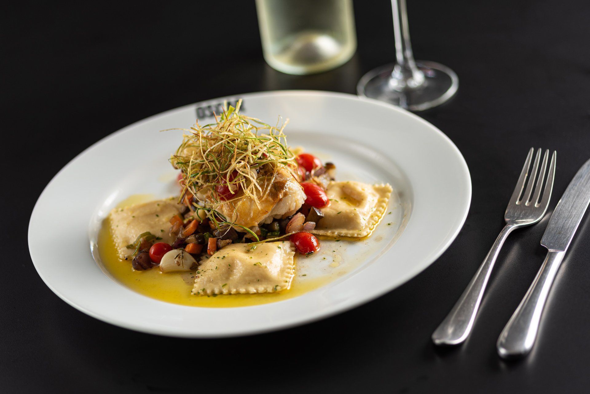Prato de ravióli com legumes e guarnição, ao lado de talheres, taça de vinho e garrafa.