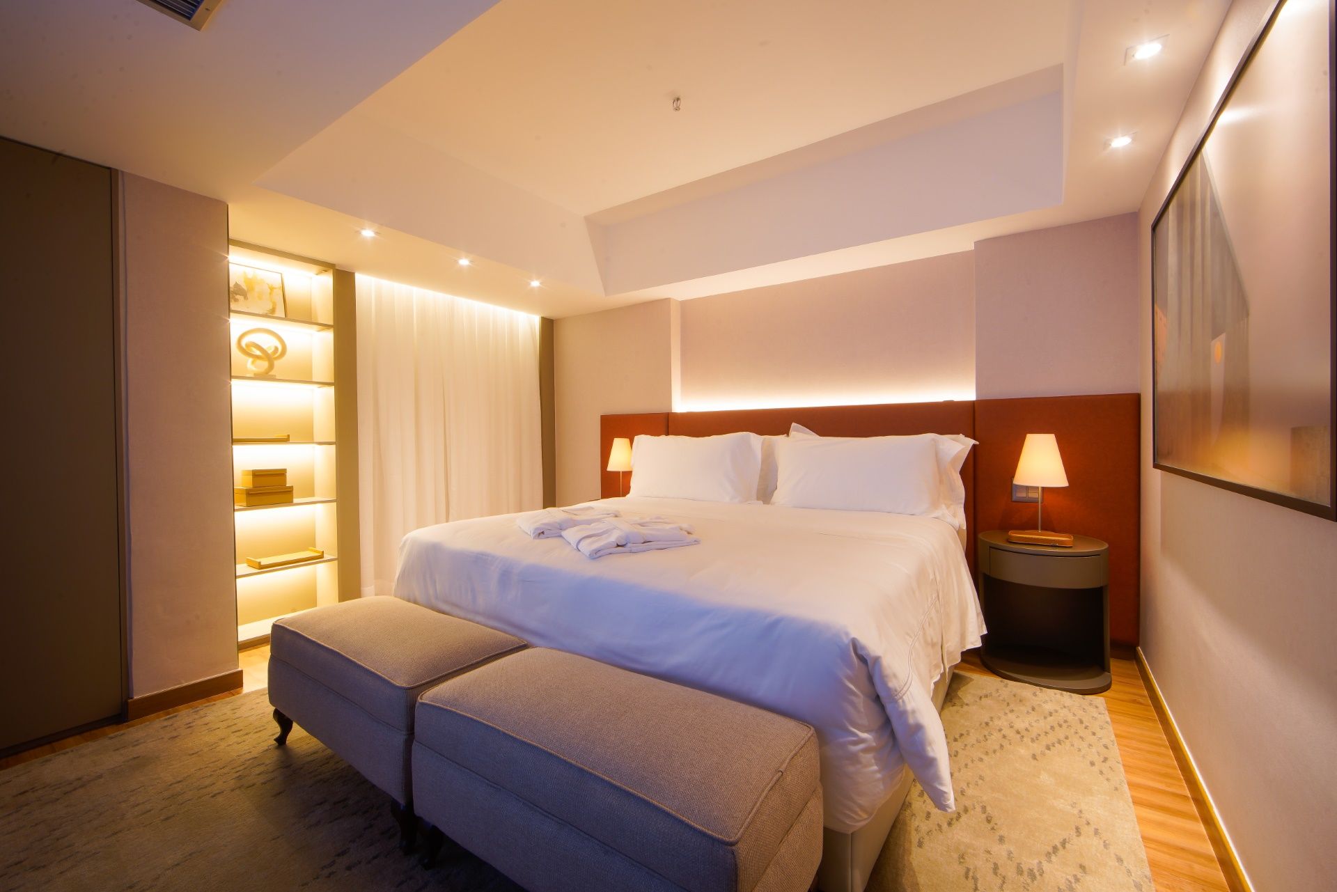 Quarto de hotel aconchegante com cama king-size, bancos, luminárias e uma estante iluminada decorativa.