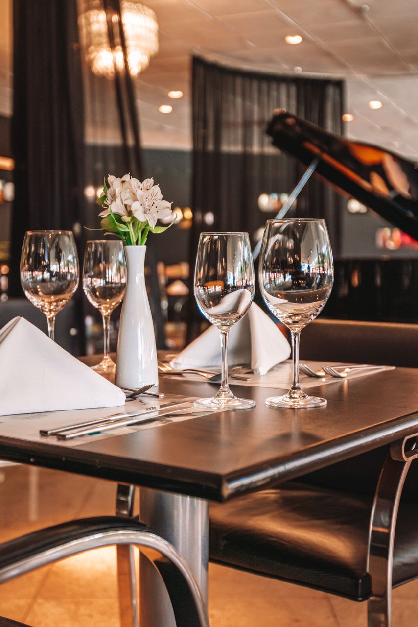 Uma mesa de restaurante posta para os clientes com copos de água, flores brancas e um piano escuro ao fundo.