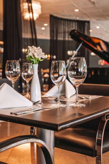 Uma mesa de restaurante posta para os clientes com copos de água, flores brancas e um piano escuro ao fundo.