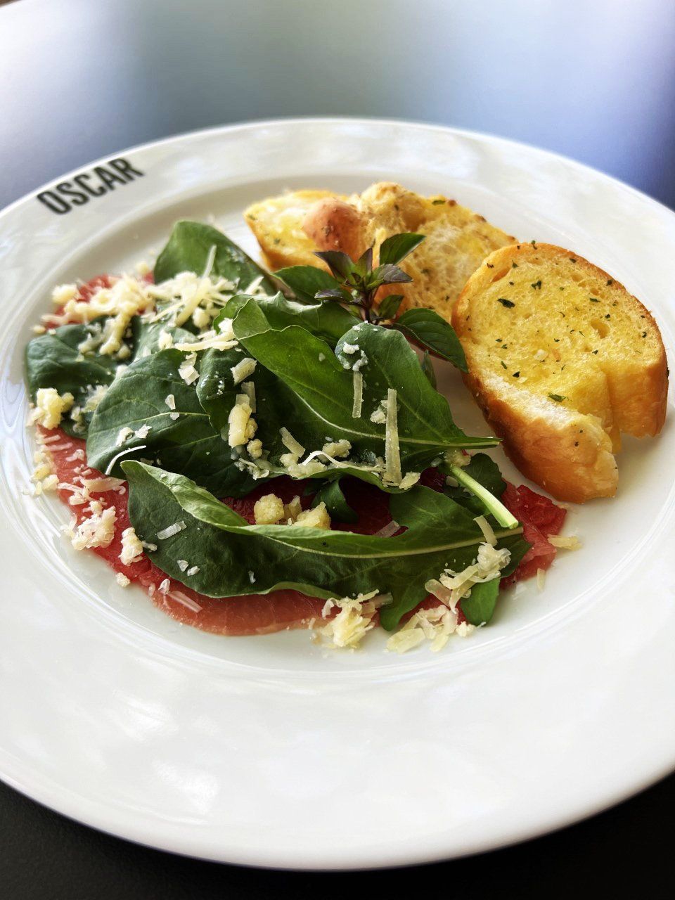 Carpaccio com rúcula e queijo, servido com pão de alho em um prato branco.