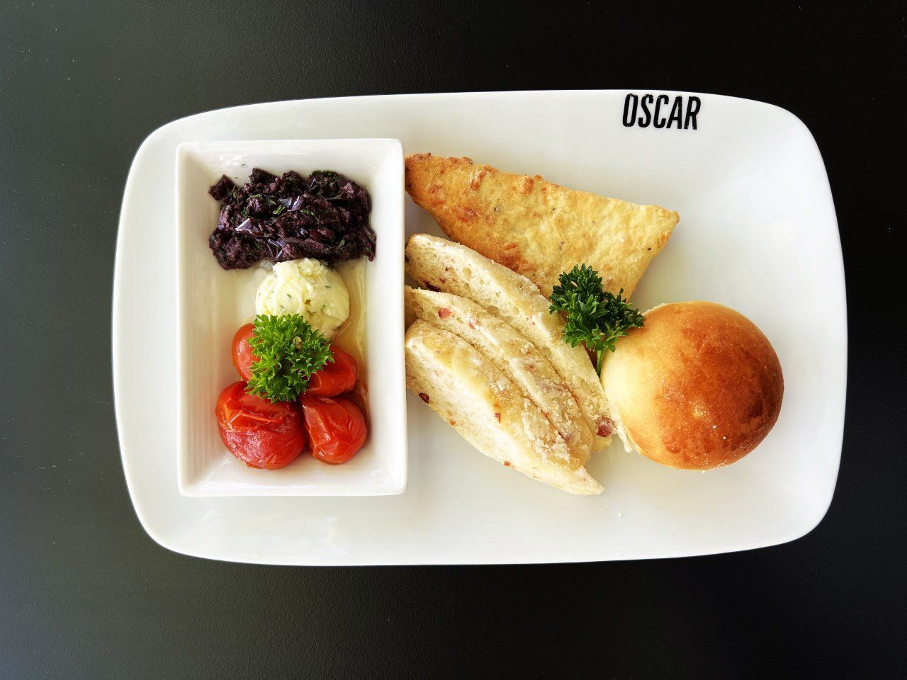 Prato de comida: pão, tomates, frutas vermelhas, manteiga, pãozinho e frango, em um prato branco sobre um fundo preto.