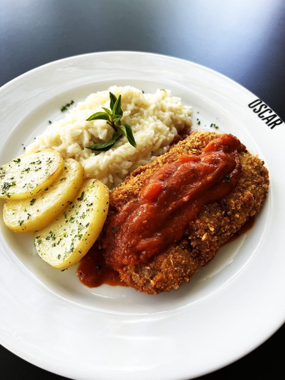 Costeleta empanada com molho de tomate, purê de arroz e batatas em um prato branco.