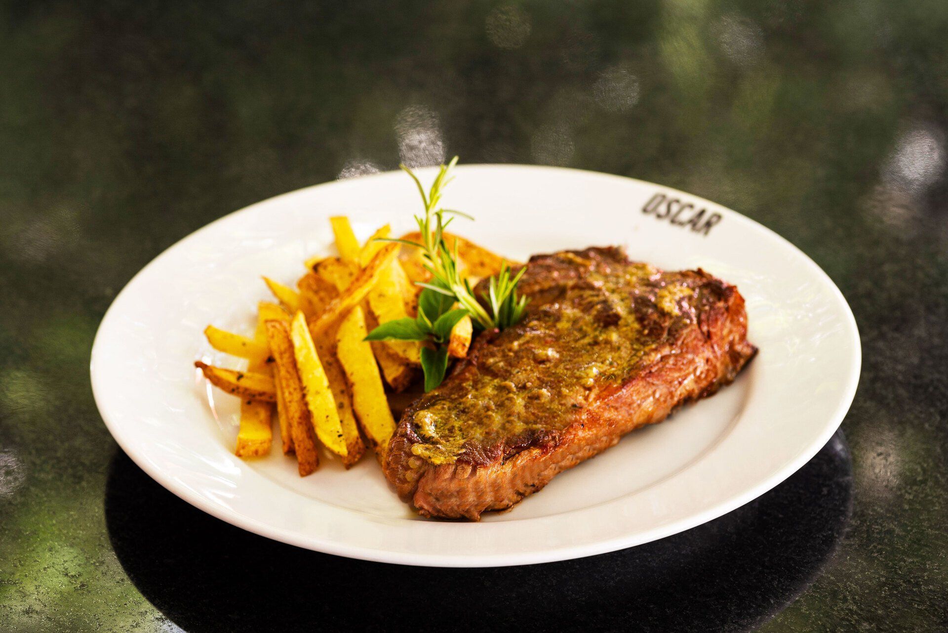 Bife com batatas fritas e alecrim em um prato branco; colocado em uma superfície escura.