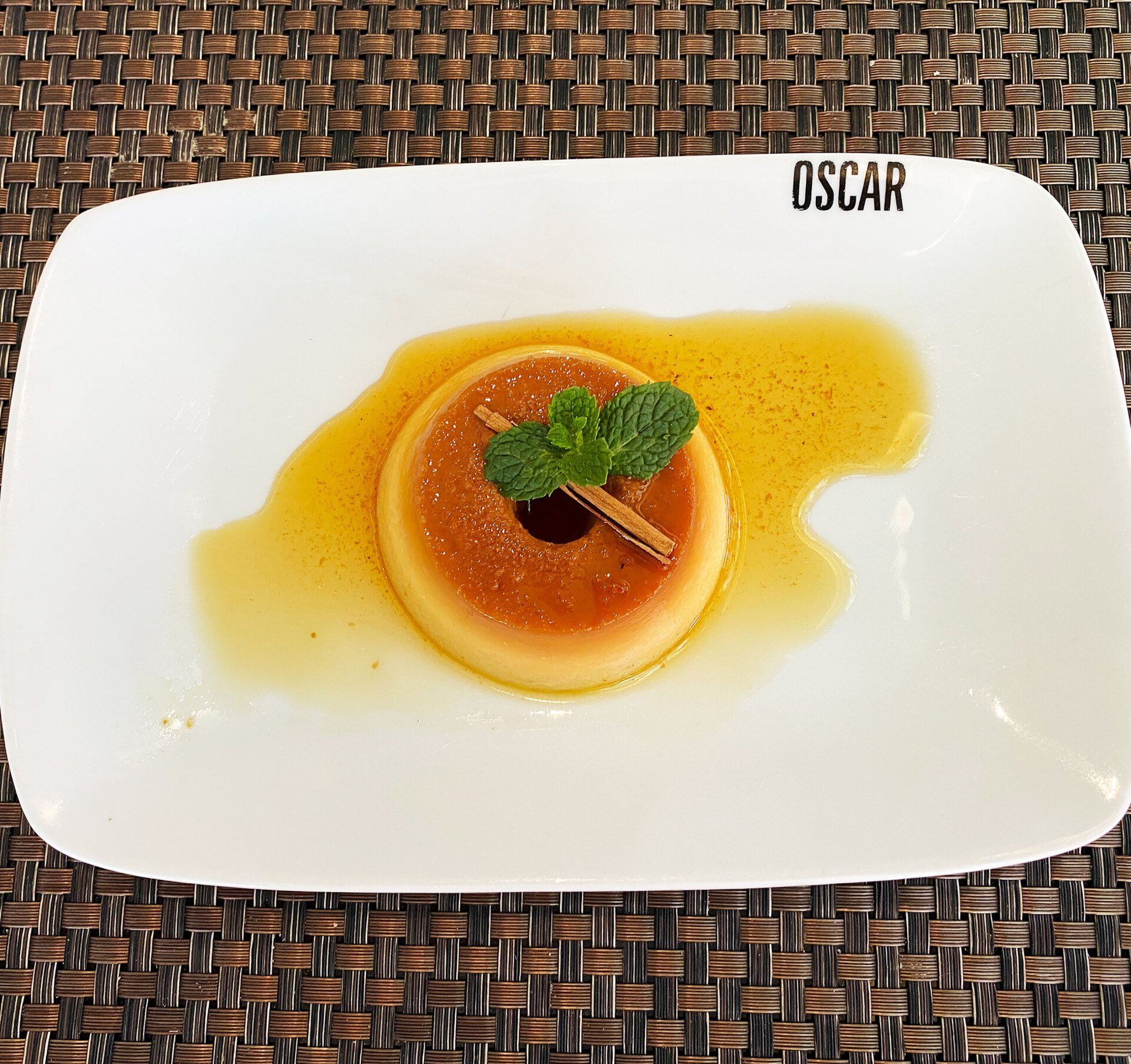 Sobremesa flan em um prato branco, regada com calda de caramelo, decorada com hortelã e um pau de canela.
