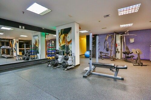 Academia com pesos, aparelhos e espelhos. Piso preto, teto branco e detalhes coloridos.