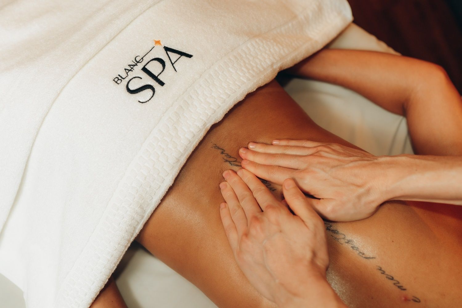 Pessoa recebendo uma massagem nas costas em um spa. Mãos exercem pressão sobre a pele oleosa. Um robe branco cobre os ombros.