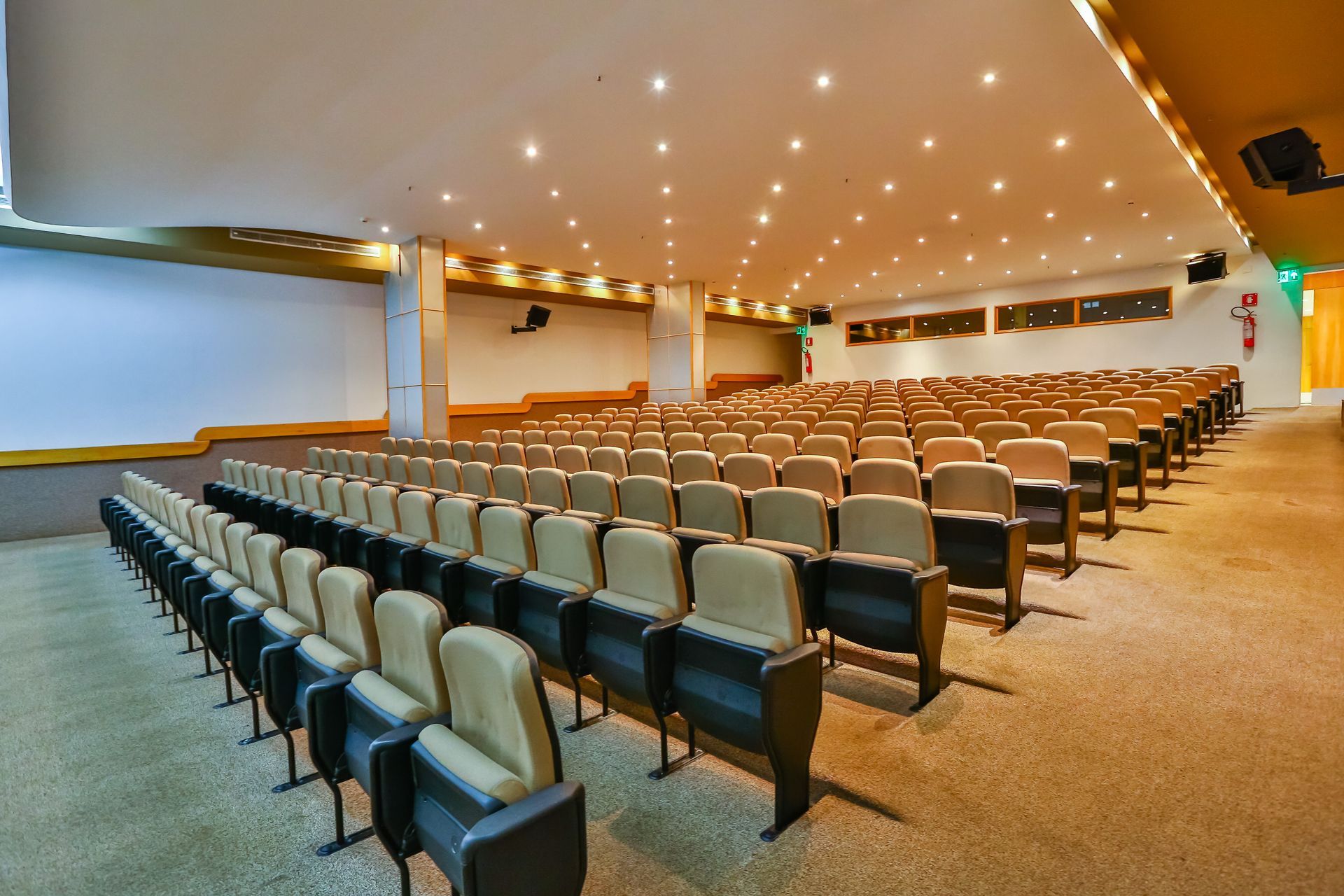 Auditório com fileiras de cadeiras bege e pretas, uma tela de projeção e luzes suspensas.