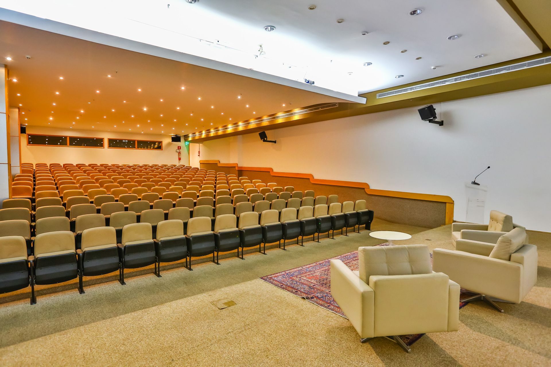 Auditório com poltronas em camadas, poltronas creme e palco. Iluminação aconchegante e cores neutras.