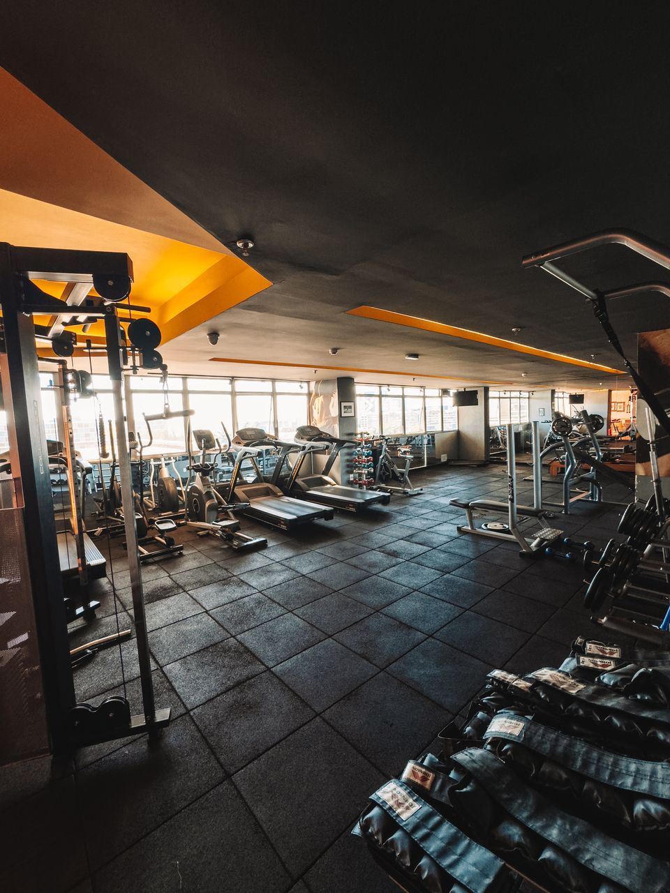 Interior de academia com equipamentos de ginástica, incluindo esteiras e aparelhos de musculação, sob um teto escuro e detalhes em amarelo brilhante.