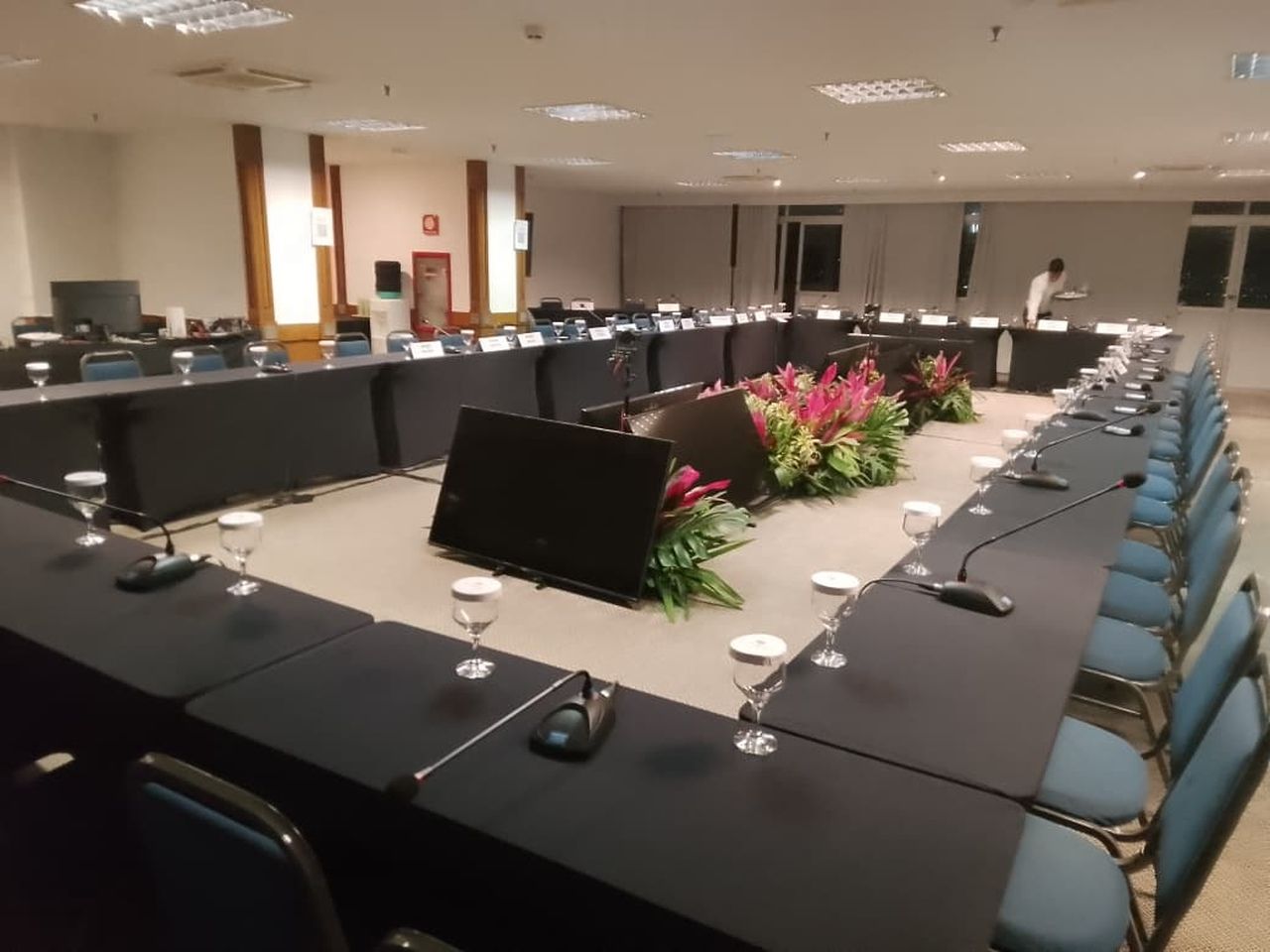 Sala de conferências com mesas em formato de U, microfones, cadeiras e um centro de mesa floral.