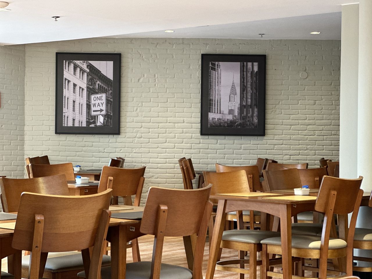 Interior do restaurante com mesas e cadeiras de madeira, parede de tijolos e fotos emolduradas em preto e branco.