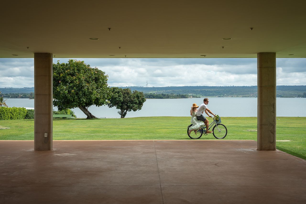 Pessoa andando de bicicleta em uma área gramada com água ao fundo, vista através dos pilares de um edifício.