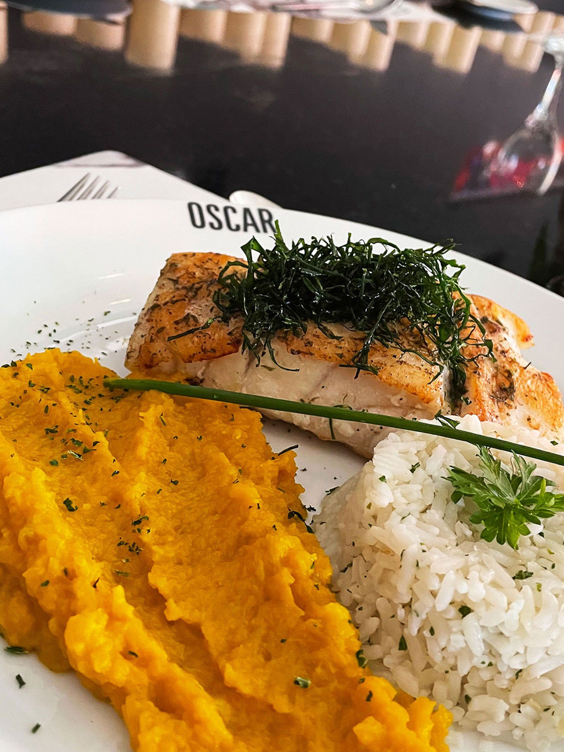 Peixe grelhado coberto com ervas, purê de batata-doce e arroz em um prato branco.