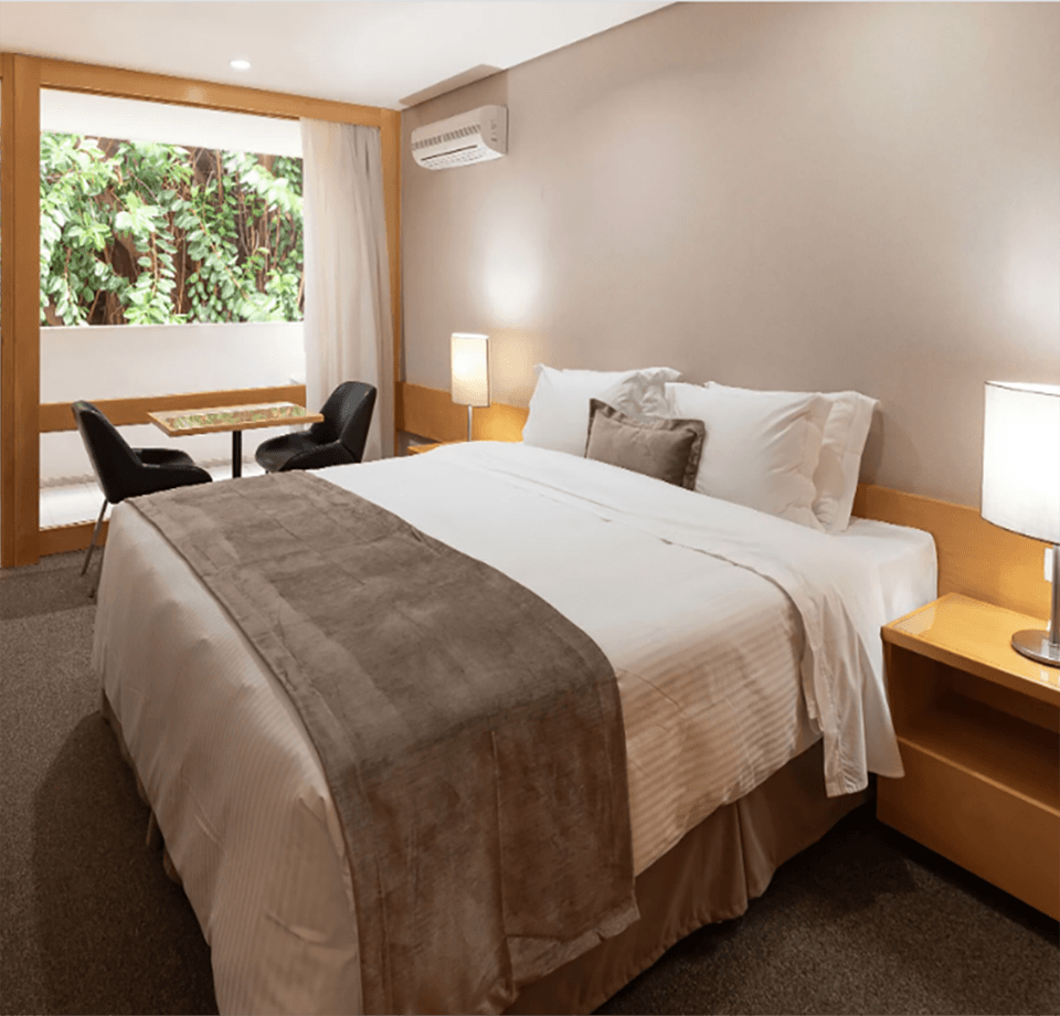 Quarto de hotel com uma cama grande, mesas de cabeceira e uma janela com vista para a área verde.