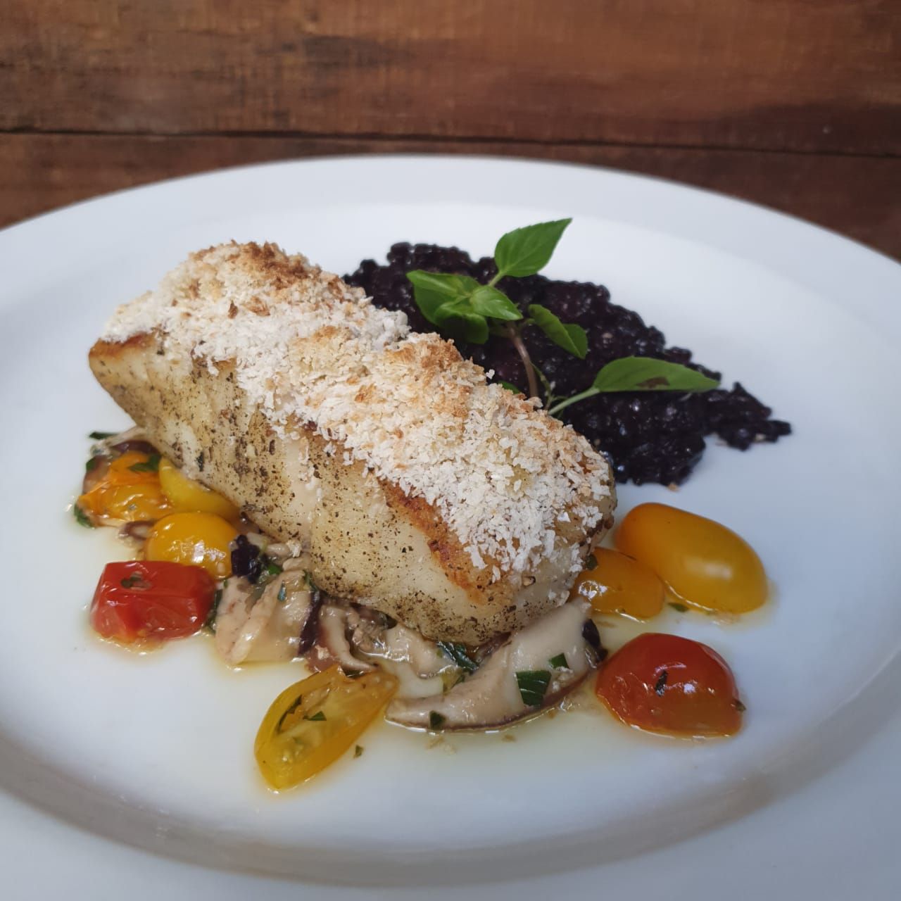 Filé de peixe com crosta sobre tomates e cogumelos salteados, com arroz preto e manjericão em um prato branco.