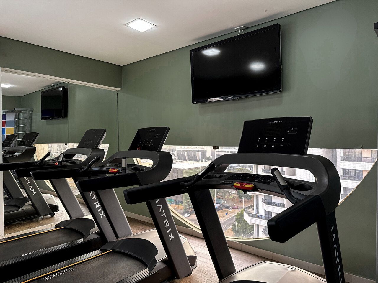 Academia com esteiras, TV na parede e espelho. O ambiente é interno, com cores neutras.