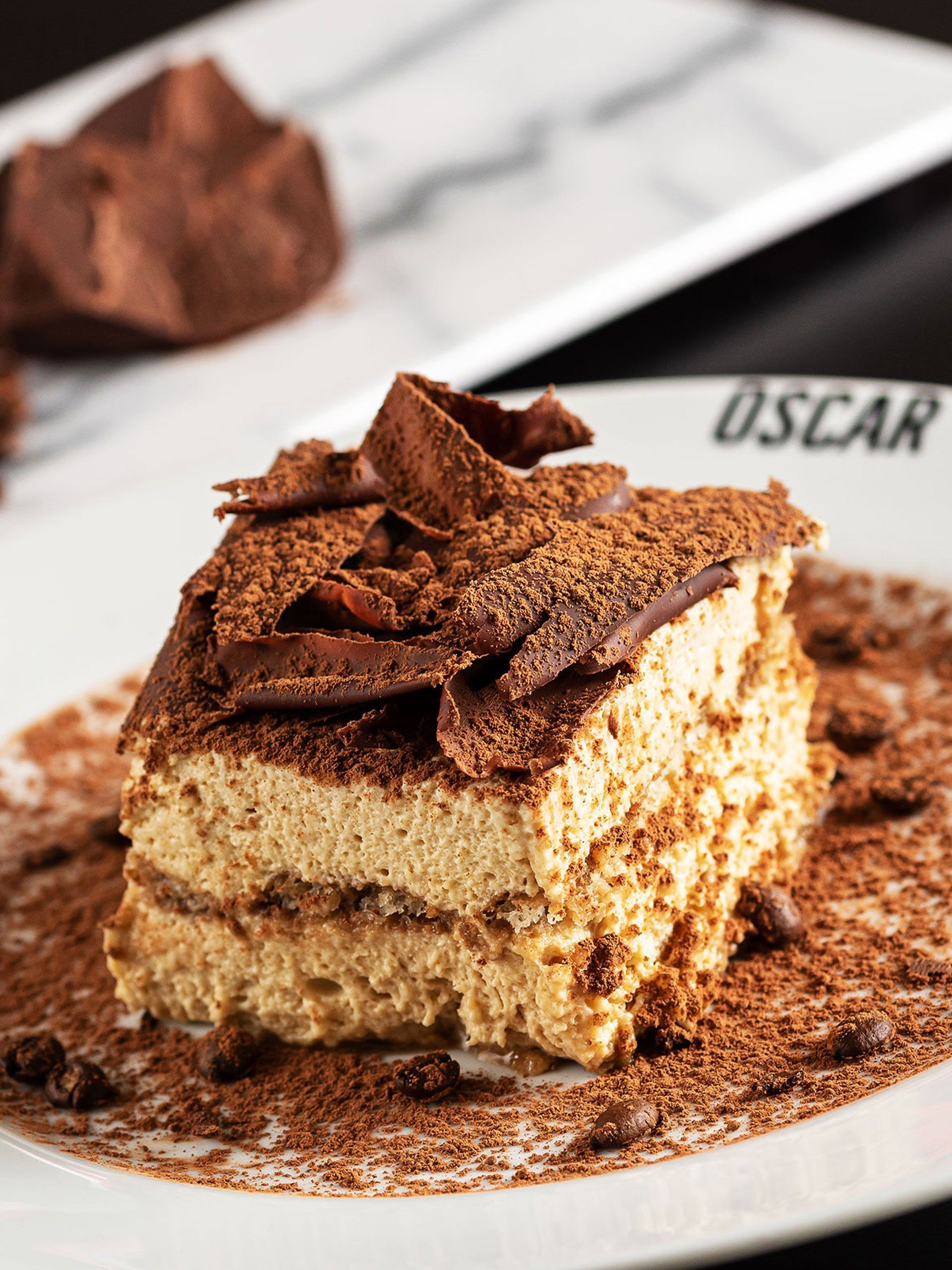 Sobremesa tiramisu em um prato branco, polvilhada com cacau em pó e raspas de chocolate.