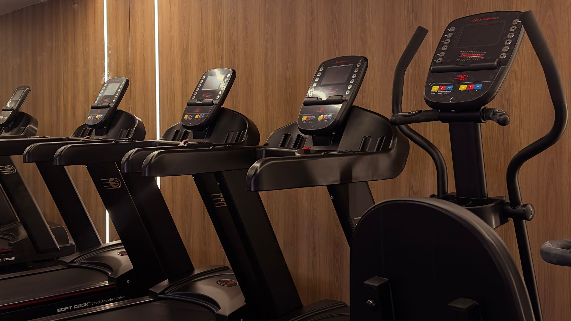 Uma fileira de aparelhos de ginástica pretos, incluindo esteiras e um elíptico, enfileirados contra uma parede de madeira.