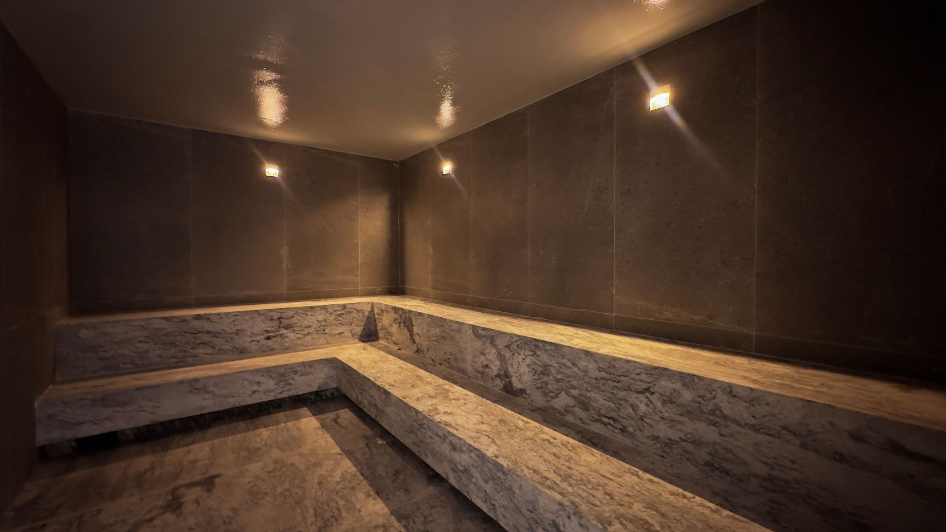 Interior de sauna moderno com assentos de pedra em níveis, paredes escuras e iluminação embutida aconchegante.