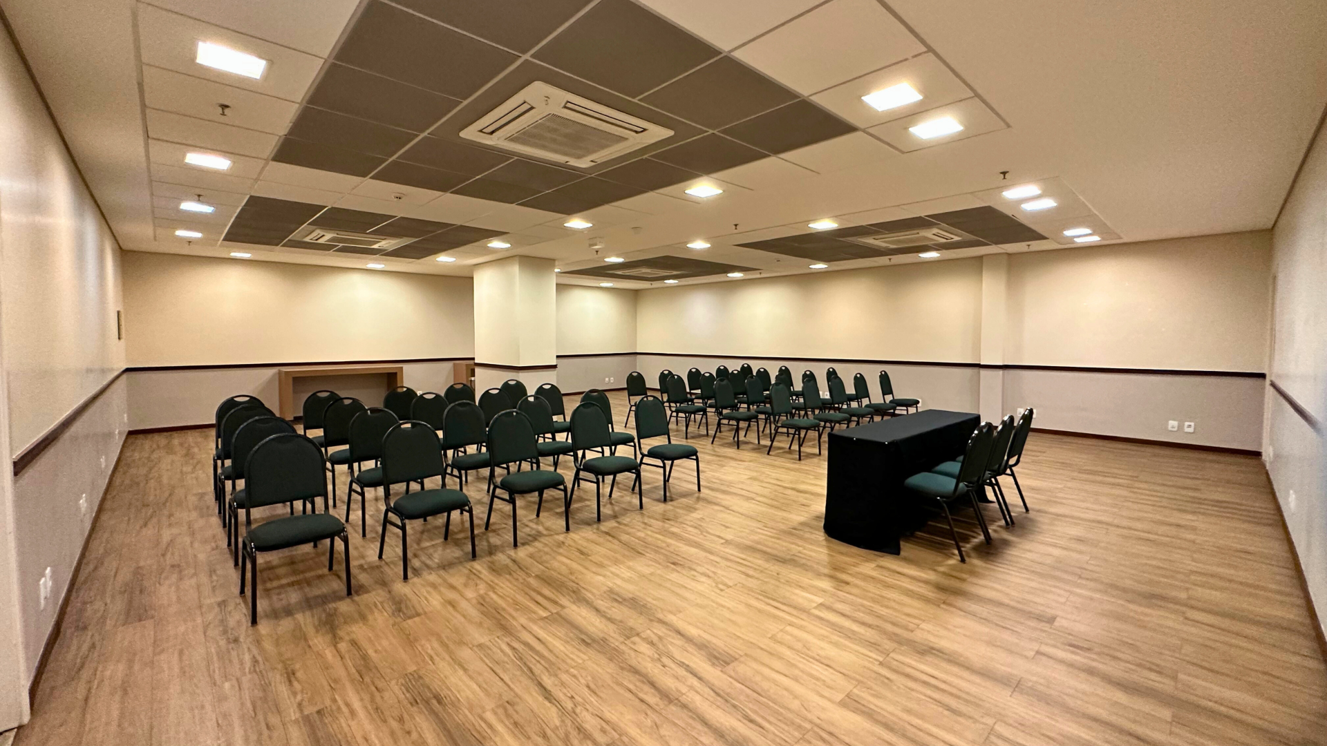 Uma sala de conferências vazia com fileiras de cadeiras voltadas para a mesa de um palestrante. Pisos de madeira e paredes neutras.
