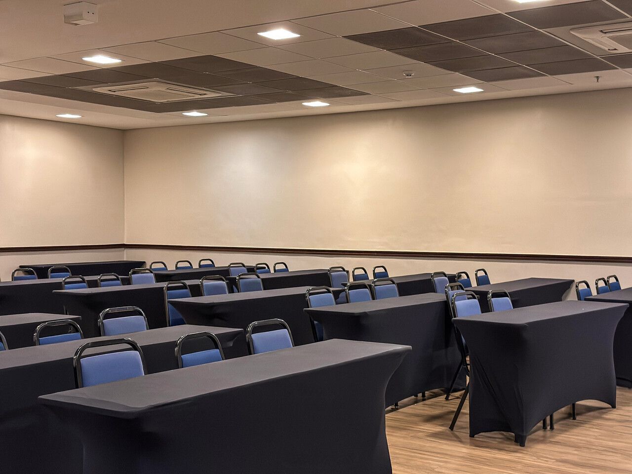 Sala de conferências vazia com fileiras de mesas cobertas com toalhas pretas e cadeiras azuis.