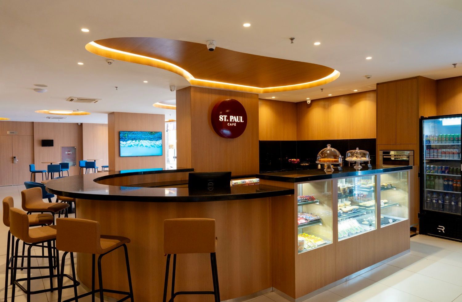 Café do hotel com balcão e assentos, acabamentos em madeira e preto, exposição de doces e refrigerador de bebidas.