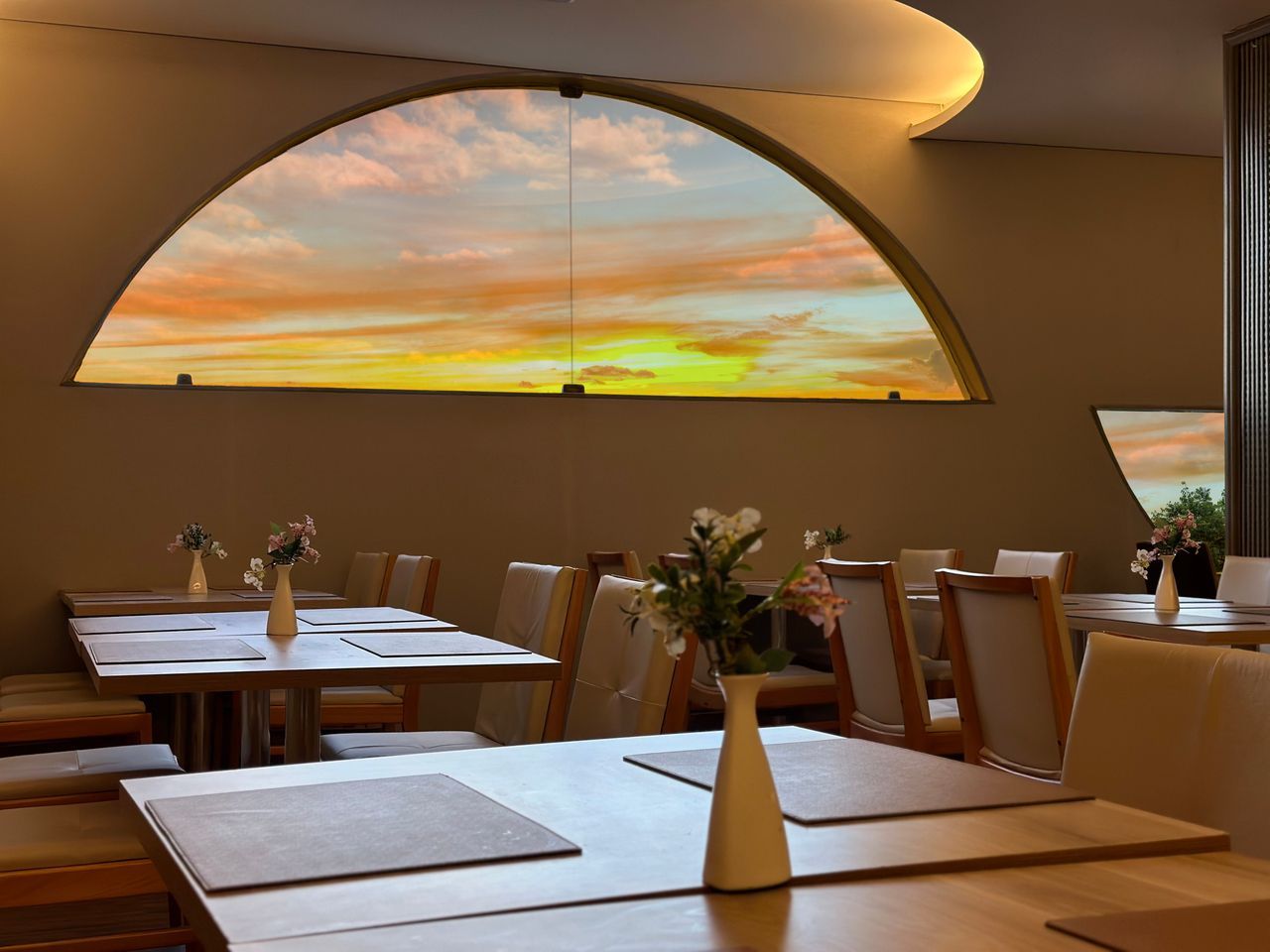 Interior do restaurante com mesas postas, com um mural do pôr do sol em uma janela em arco.