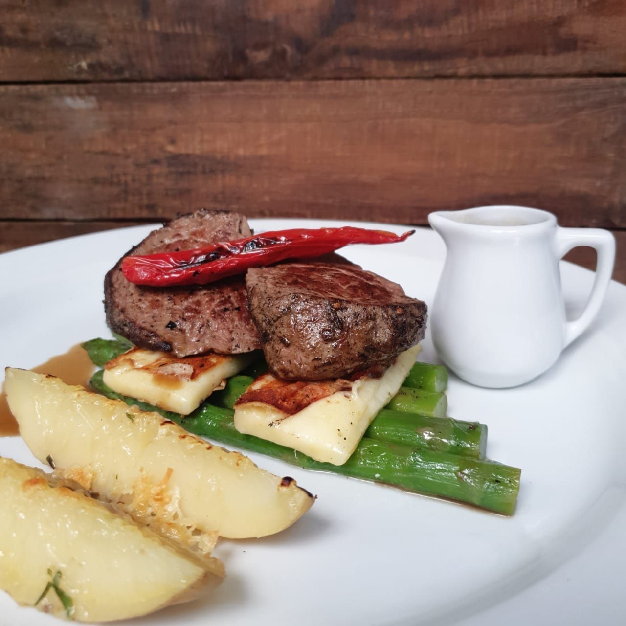 Prato de bife com vegetais: bife, halloumi, aspargos, batatas, pimenta, molho em uma jarra pequena.