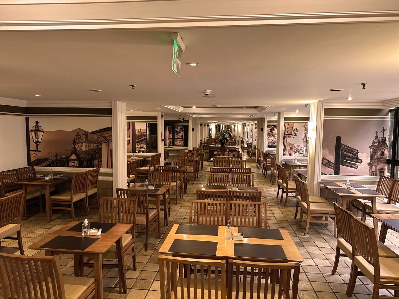 Interior do restaurante com mesas e cadeiras de madeira, murais de paisagens e piso de cerâmica.