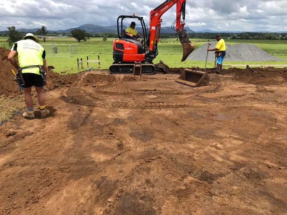 A Man Is Standing Next To An Excavator In A Dirt Field — Luke Seears Concreting in Laurieton, NSW